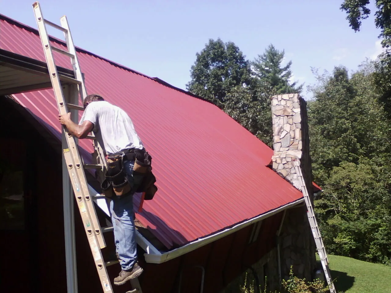 Roofer on a ladder beside a red metal roof and stone chimney — Soffit & Fascia Repair by Roofing Brothers in Stevensburg